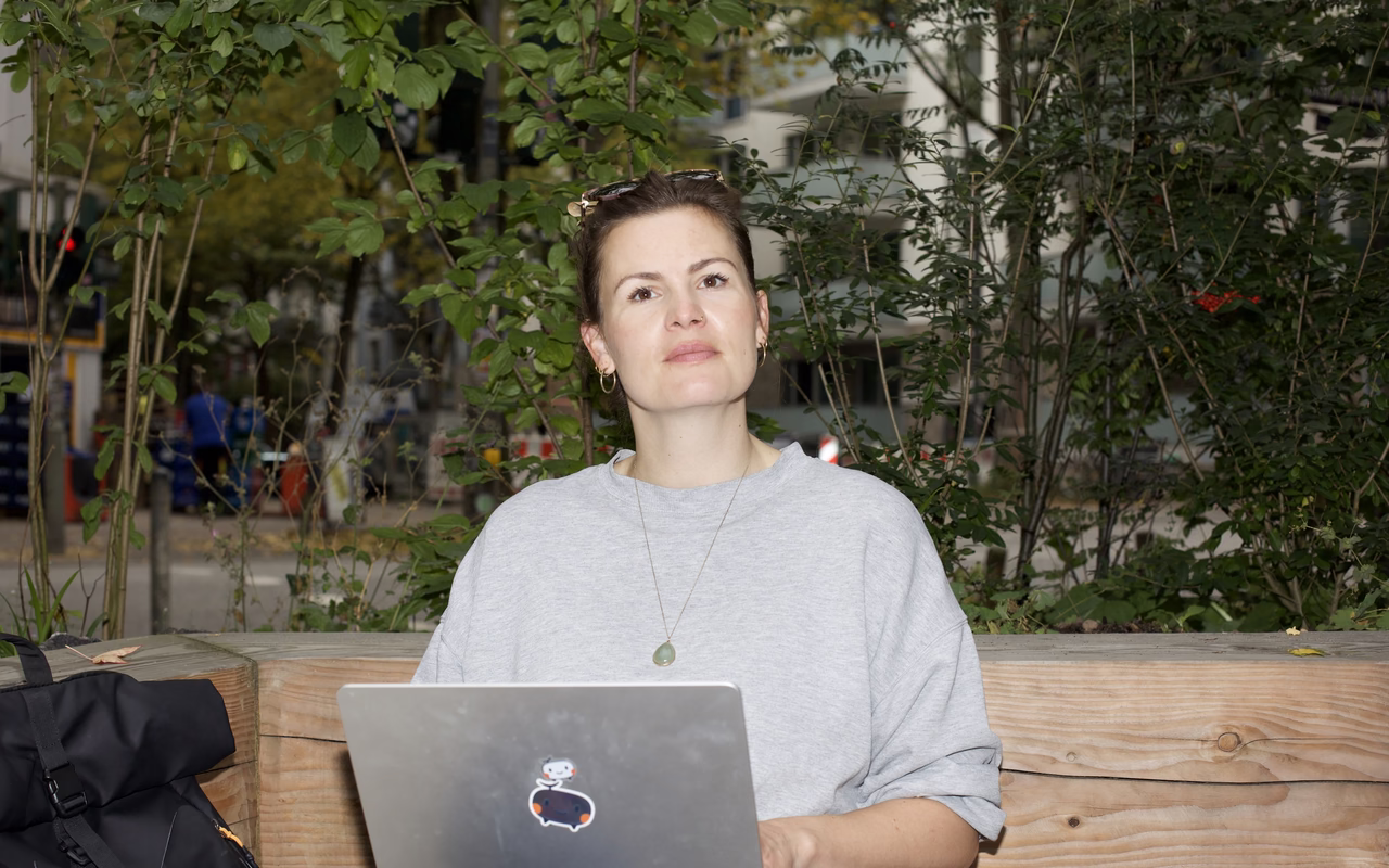 Melanie Wildt sitzt mit Laptop auf einer Holzbank zwischen Sträuchern und blickt in die Kamera.