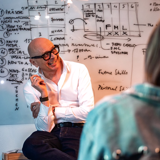 Marcus John Henry Brown sitzt lachend vor einem beschriebenen Whiteboard, im Vordergrund unscharf eine Zuhörerin.