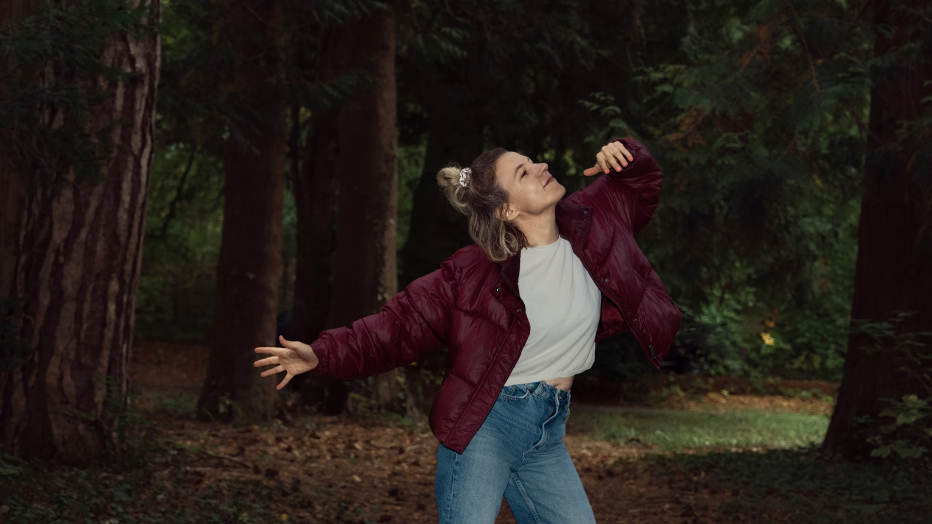 Alena Wollert steht mit ausgebreiteten Armen und geschlossenen Augen zwischen hohen Bäumen im Wald, sie trägt eine dunkelrote Jacke.