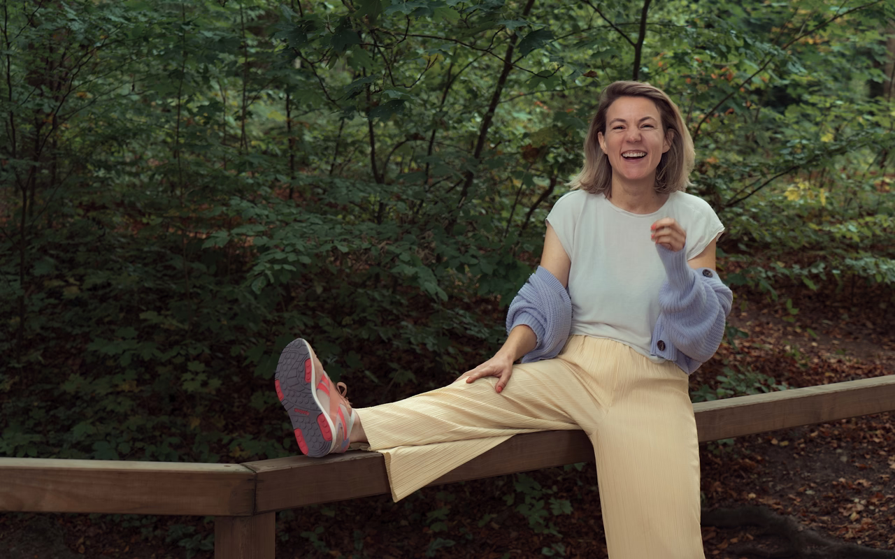 Alena Wollert sitzt lachend auf einem Holzgeländer im Wald und streckt ein Bein darauf aus.