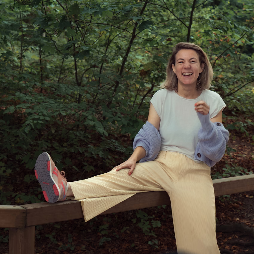 Alena Wollert lacht auf einem Holzgeländer im Wald, eine fliederfarbene Strickjacke hängt ihr über die Arme.