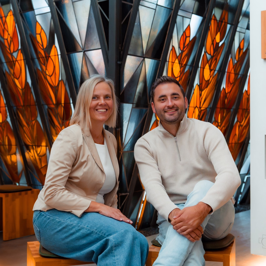 Tanja Bosbach und Tim Busam sitzen auf Holzhockern vor einem orange leuchtenden Glasfenster.