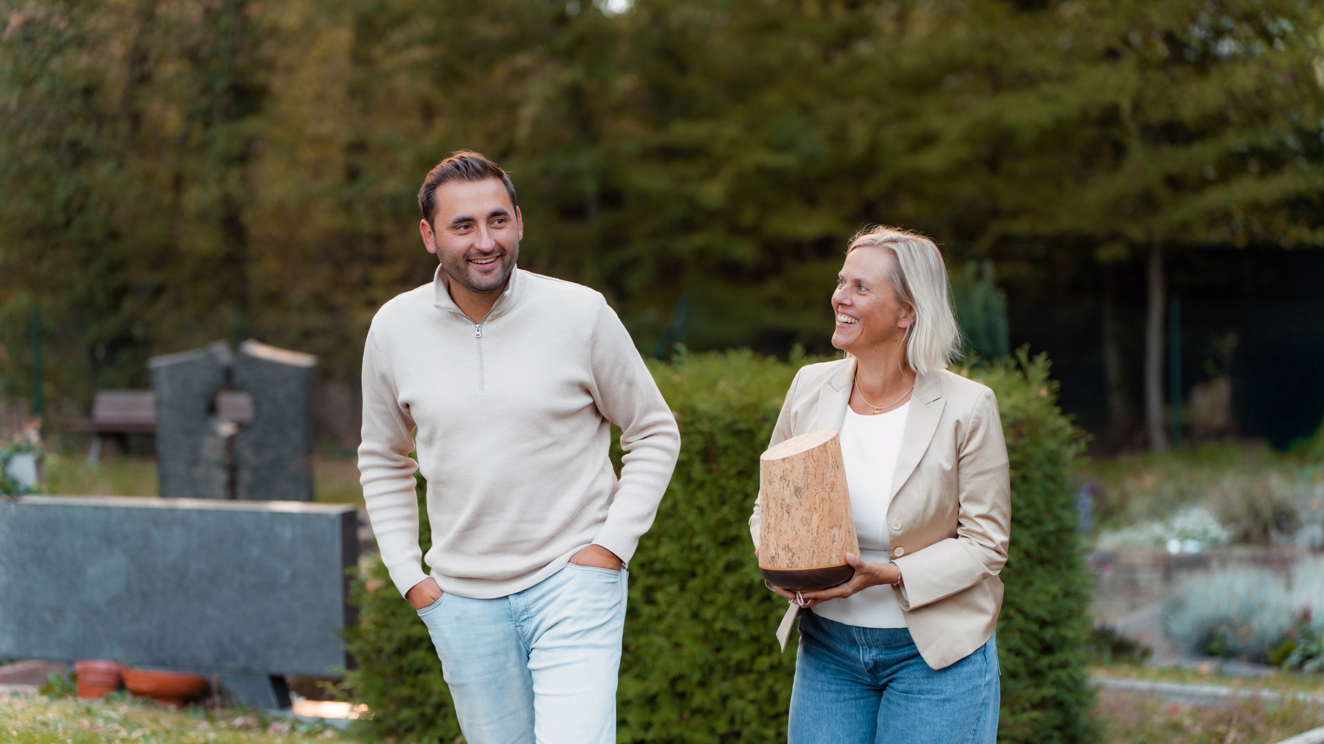 Tim Busam und Tanja Bosbach gehen lachend über einen Friedhof, Tanja trägt eine helle Urne.
