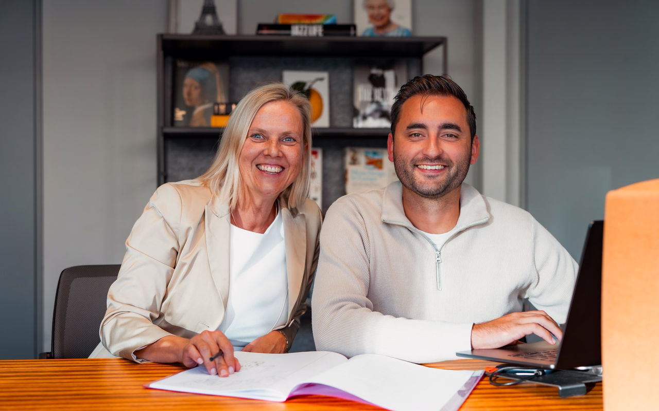Tanja Bosbach und Tim Busam sitzen lachend an einem Holztisch mit aufgeschlagenem Notizbuch und Laptop.