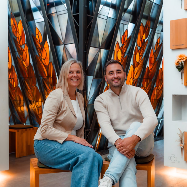 Tanja Bosbach und Tim Busam lächeln in einer Kapelle, hinter ihnen ein großes Kirchenfenster in Orange und Grau.