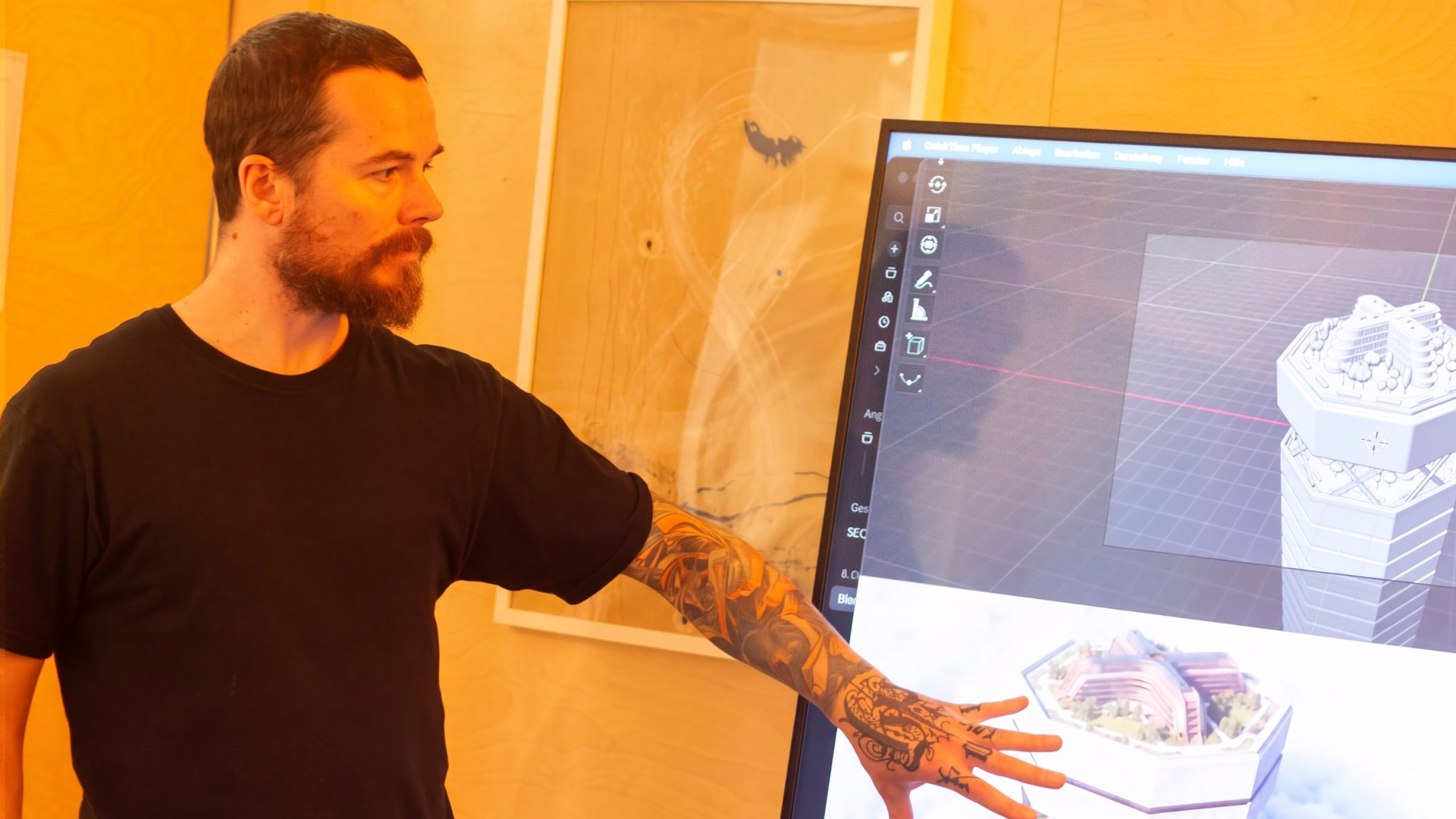 Mann im schwarzen T-Shirt zeigt auf einen Bildschirm mit dem 3D-Modell eines Gebäudes.