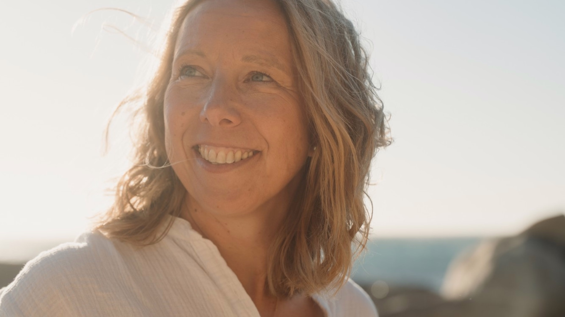Anja Helming steht am Meer und lacht, im Gegenlicht der tiefstehenden Sonne.
