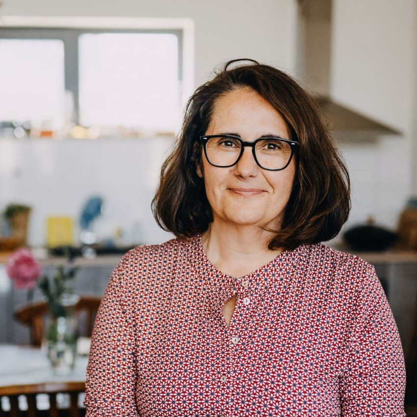 Ana Martinez Garcia mit Brille und gemusterter Bluse lehnt lächelnd an einer Sofalehne, hinter ihr eine helle Küche.