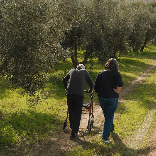 Ana Martinez Garcia geht neben ihrem Vater, der einen Rollator schiebt, auf einem Feldweg durch den Olivenhain.