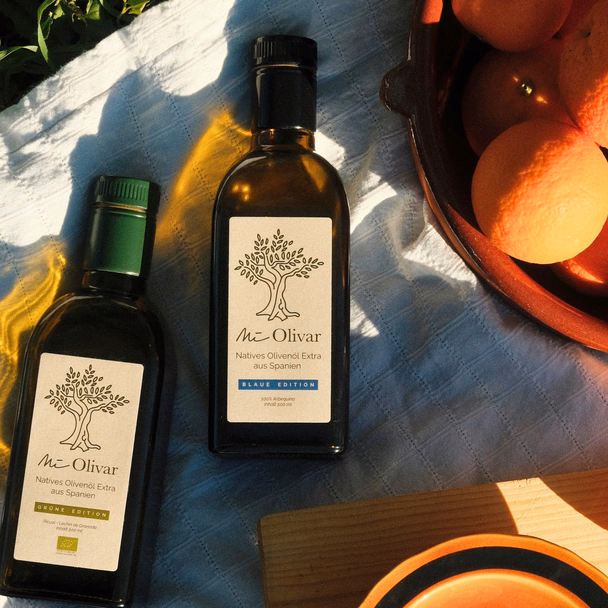 Zwei Flaschen Mi Olivar, Grüne und Blaue Edition, liegen auf einem hellen Tuch neben einer Schale mit Orangen.