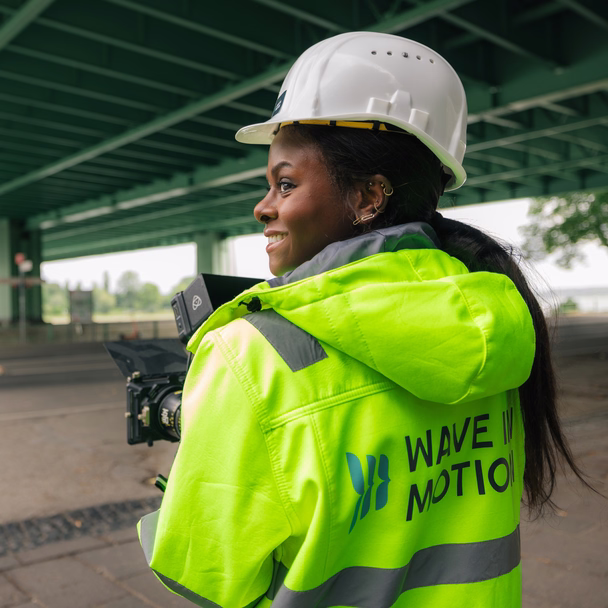 Theodora Shandé mit Schutzhelm und Warnjacke mit Wave-In-Motion-Logo blickt unter einer Brücke lächelnd zur Seite.