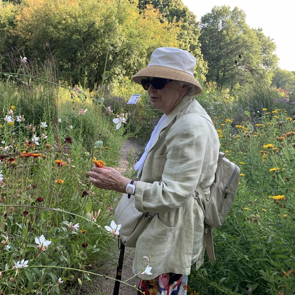 Eine ältere Dame mit Sonnenhut und Sonnenbrille betrachtet Blumen in einem blühenden Garten.
