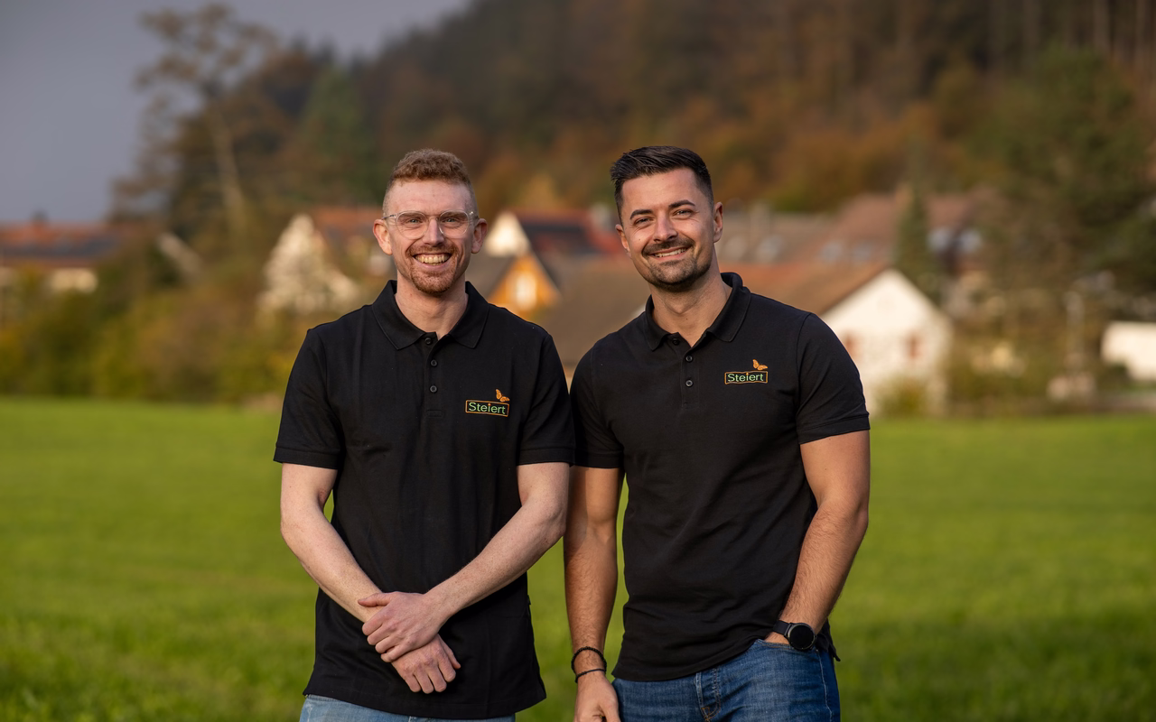 Dennis Vonderstraß und Philipp Steiert auf einer Wiese, im Hintergrund ein Dorf vor bewaldeten Hängen.