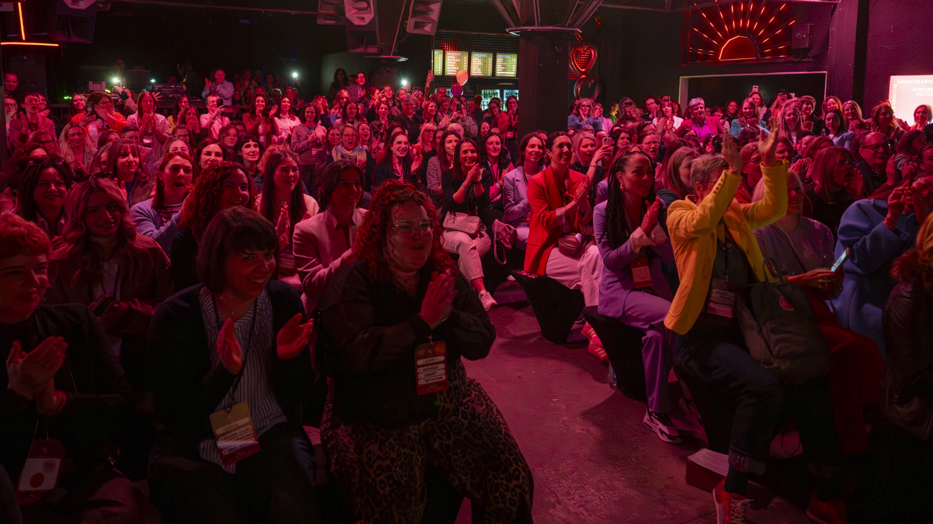 Publikum des About Us Festivals in pinkem Licht: Reihen von Frauen mit Namensschildern applaudieren und lachen.