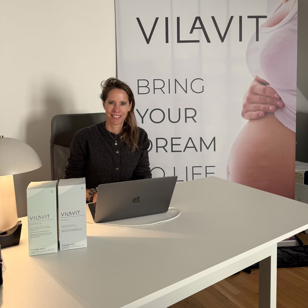 Claudia Gessler-Zwickl am Schreibtisch mit Laptop und zwei VILAVIT-Packungen, im Hintergrund ein Banner der Marke.