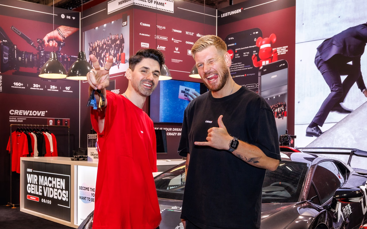 Robin Maeter im schwarzen Shirt und ein Mann im roten Shirt lachen am Messestand von CREW10, dahinter ein Lamborghini.