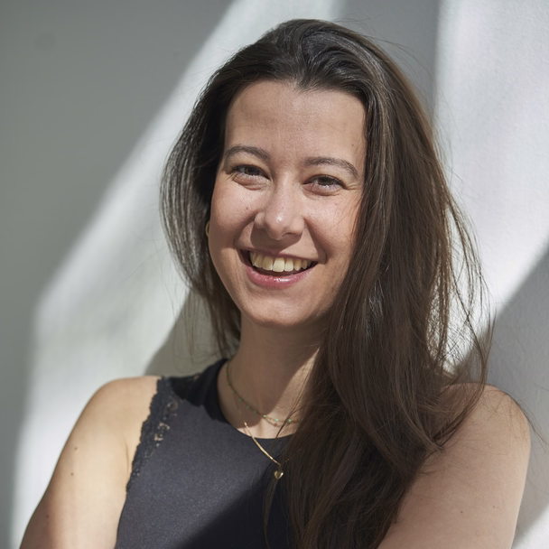 Pheline Huber lacht, an eine helle Wand gelehnt, das Haar fällt über ihre Schulter.