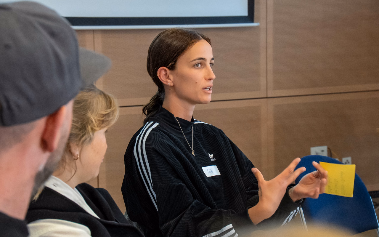Pia Wagner erklärt in einem Workshopraum etwas und gestikuliert dabei, vor ihr sitzen zwei Teilnehmende.