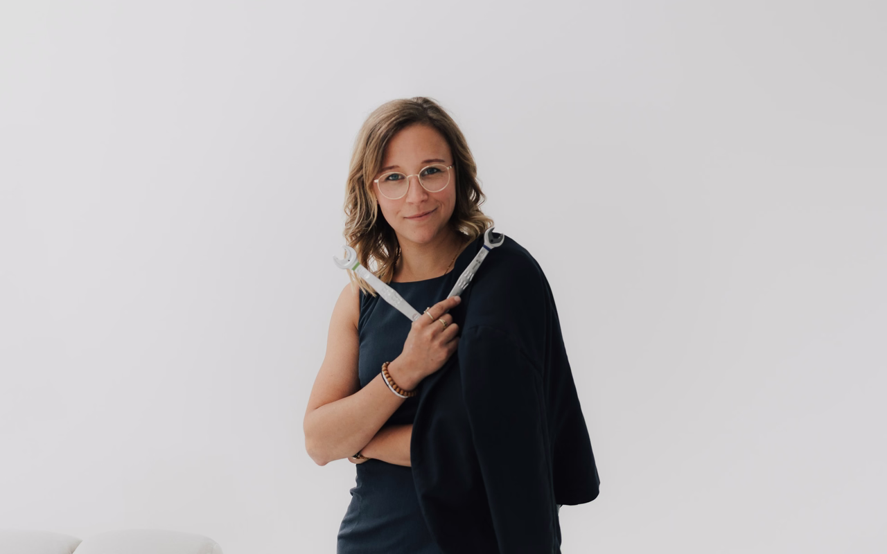 Janine Lerchen mit einer Jacke über der Schulter vor hellem Studiohintergrund.