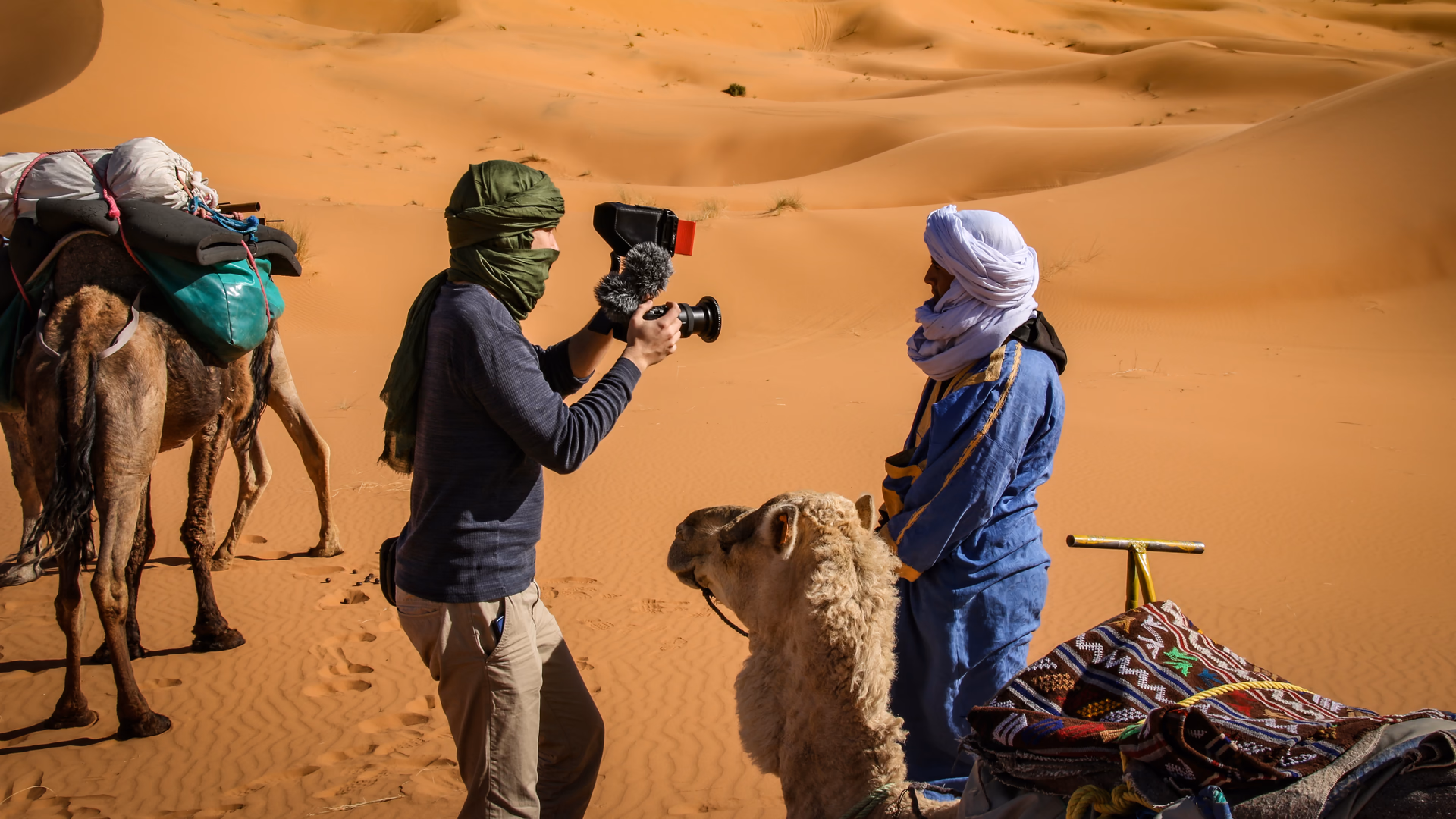 Andreas Abb filmt in der Wüste Marokkos einen Mann in blauer Tuareg-Kleidung neben einem Kamel vor Sanddünen.