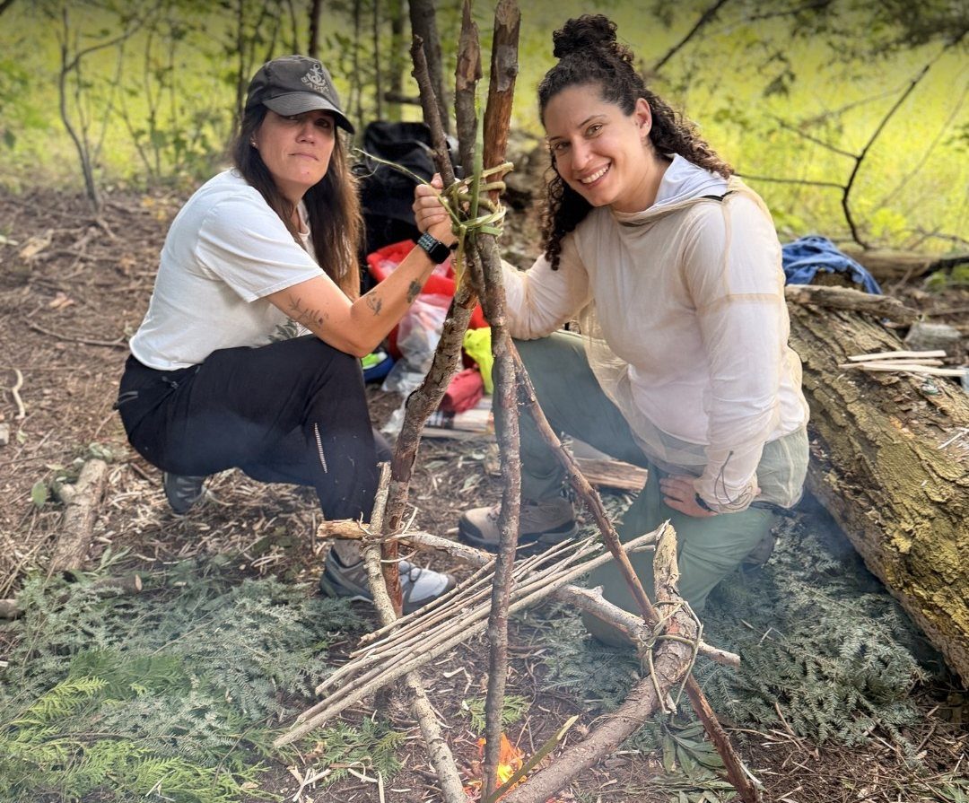 Two course participants beside a fire that has just caught under a lashed tripod