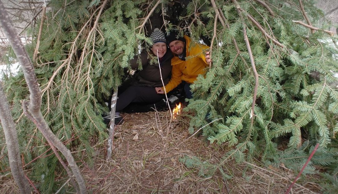 Two people sitting inside a shelter built from branches and spruce boughs