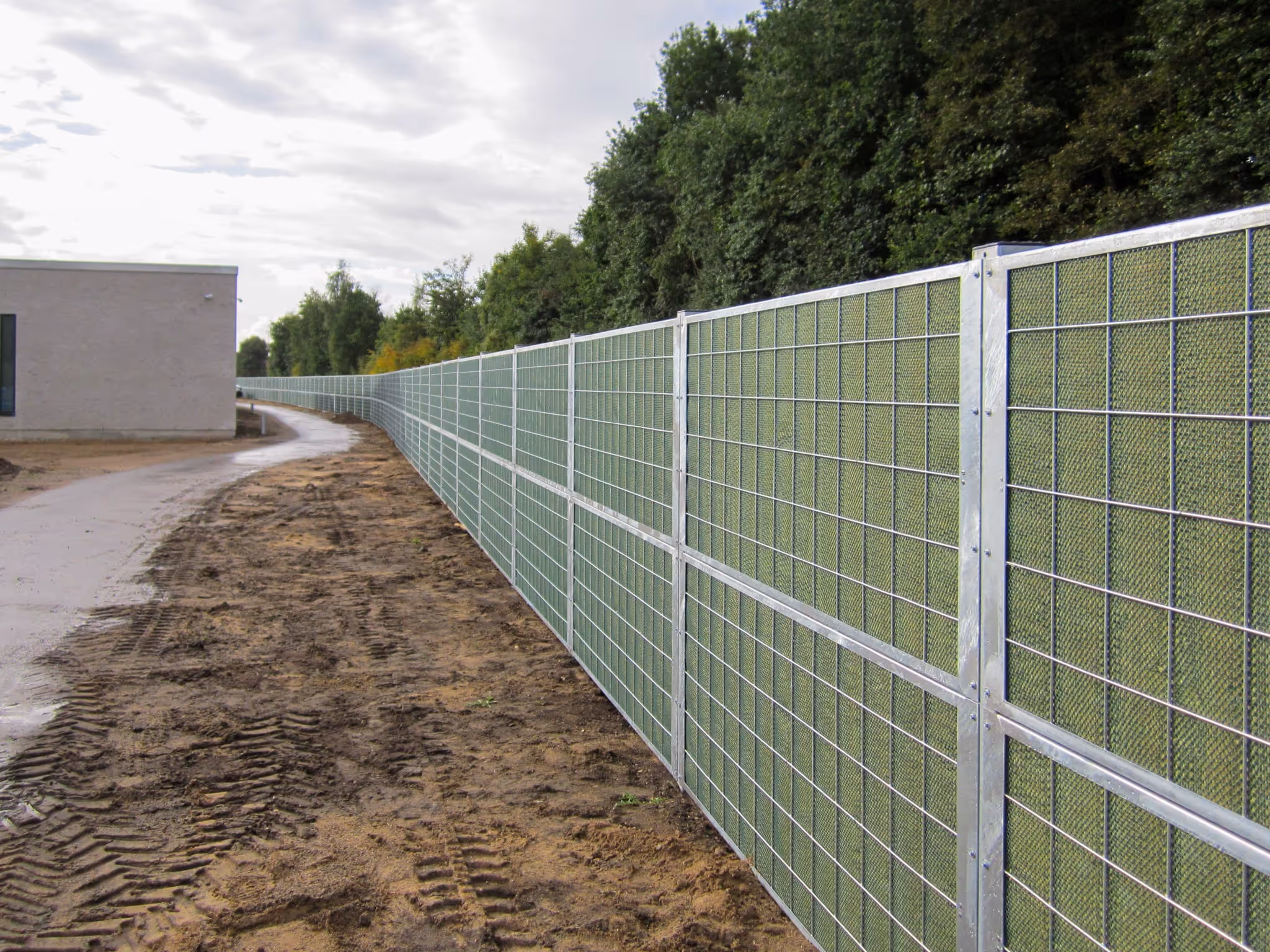 Long acoustic fence with green mesh panels along a dirt path next to dense trees and a building.