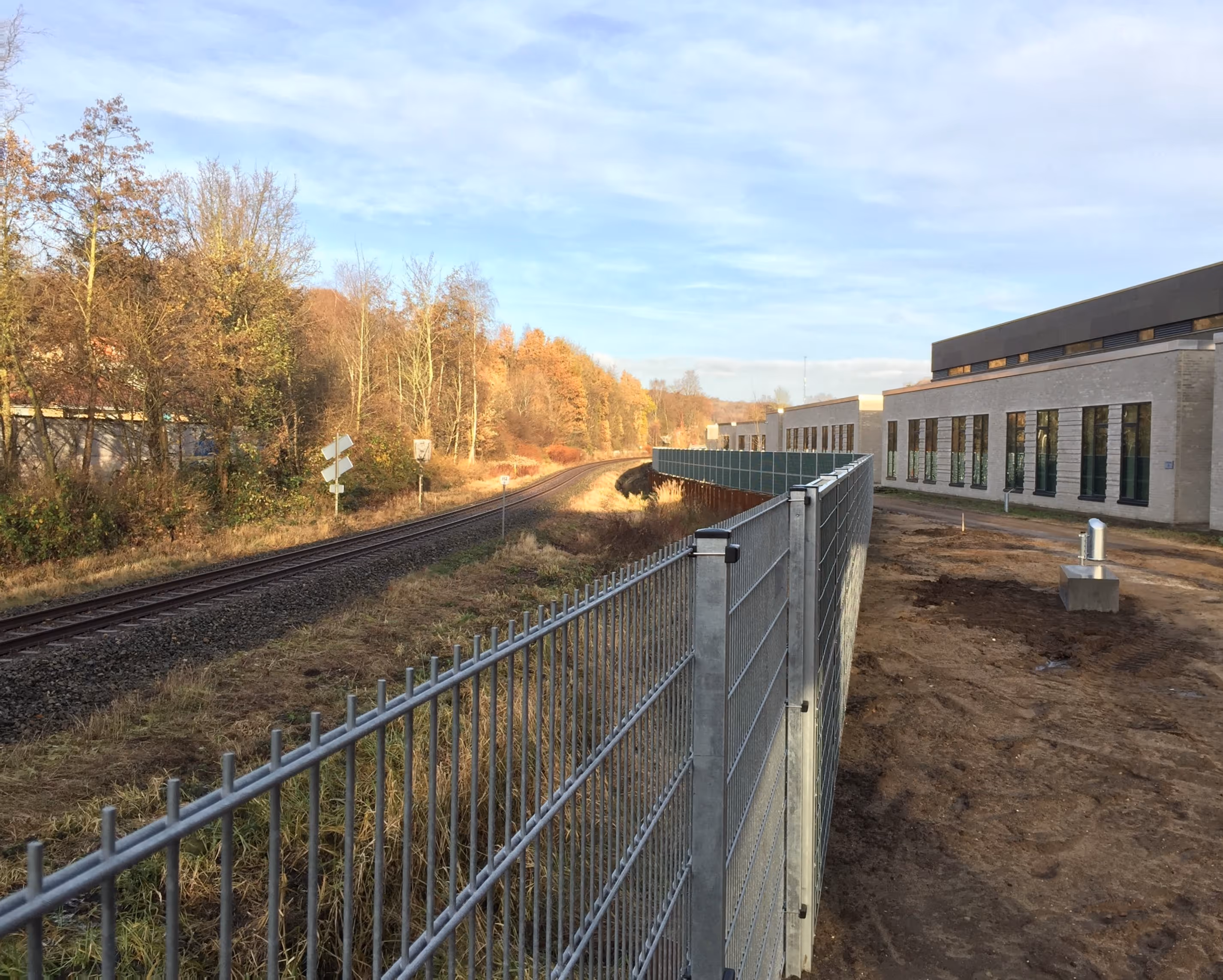 Metal fence separating a railway track and a modern building with large windows under a cloudy sky.
