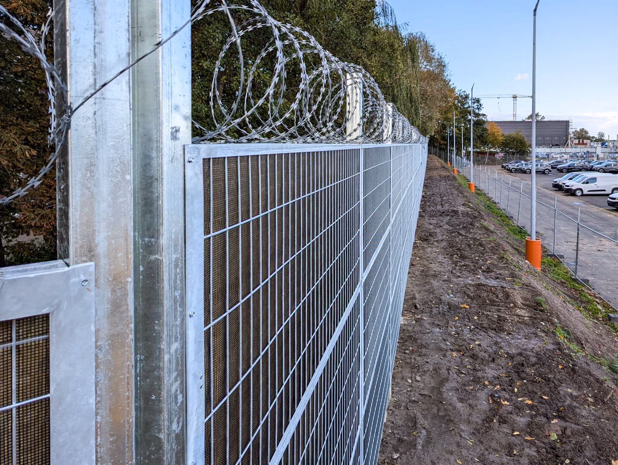 Galvanized steel security fence topped with coils of barbed wire next to a muddy path and parking lot.