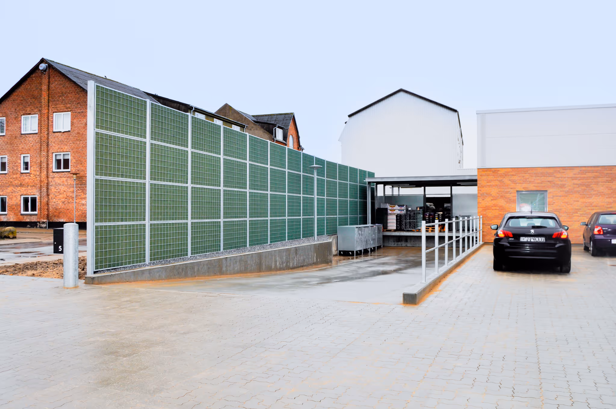 Supermarket loading area with green noise barrier walls, two parked cars, and a brick building.