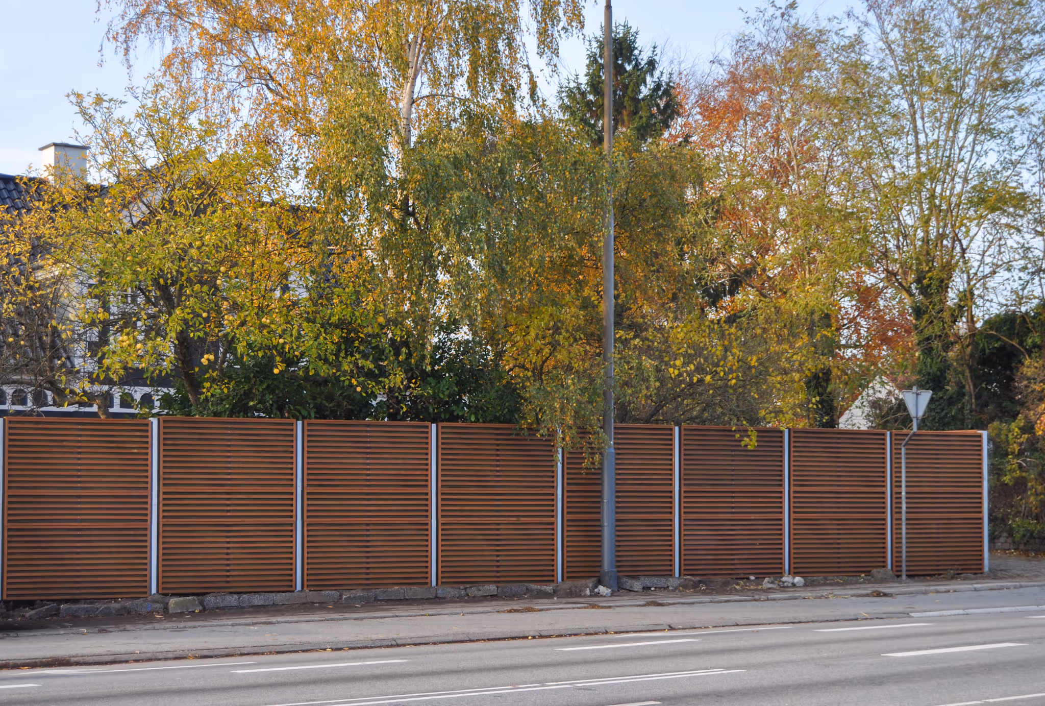 Brown slatted wooden fence along a street with autumn trees and houses in the background.