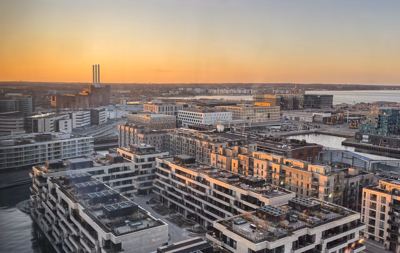 Modern cityscape at sunset with buildings, waterways, and industrial chimneys in the background.