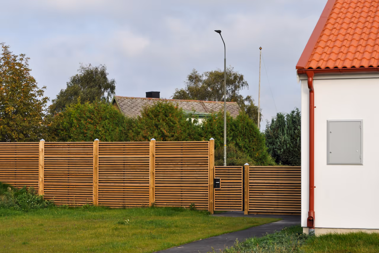 Wooden slat acoustic fence and gate beside a white house with a red tile roof and green lawn.