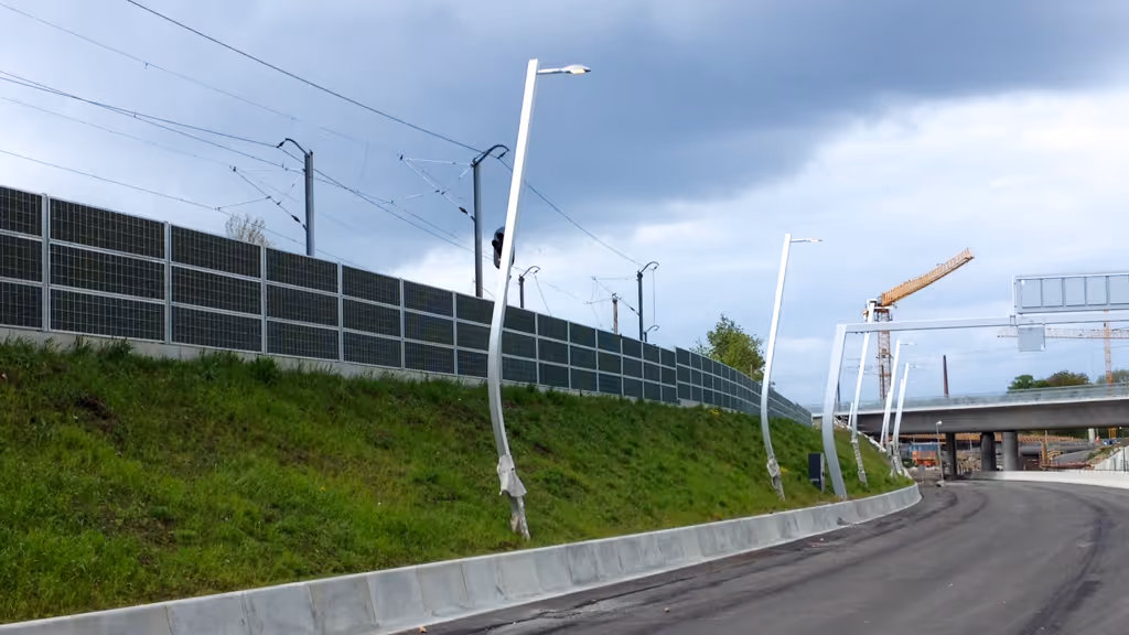 Road under construction with bent streetlights and a sound barrier beside grassy embankment.