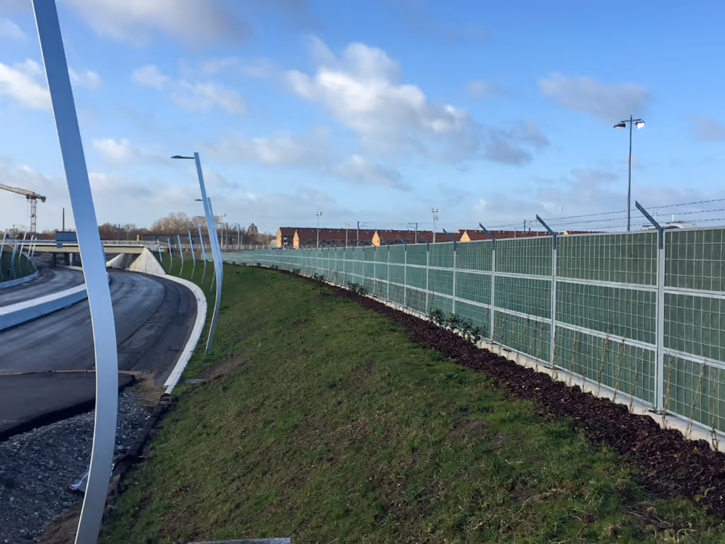 Curved road beside a green acoustic steel fence and grass under a partly cloudy blue sky.