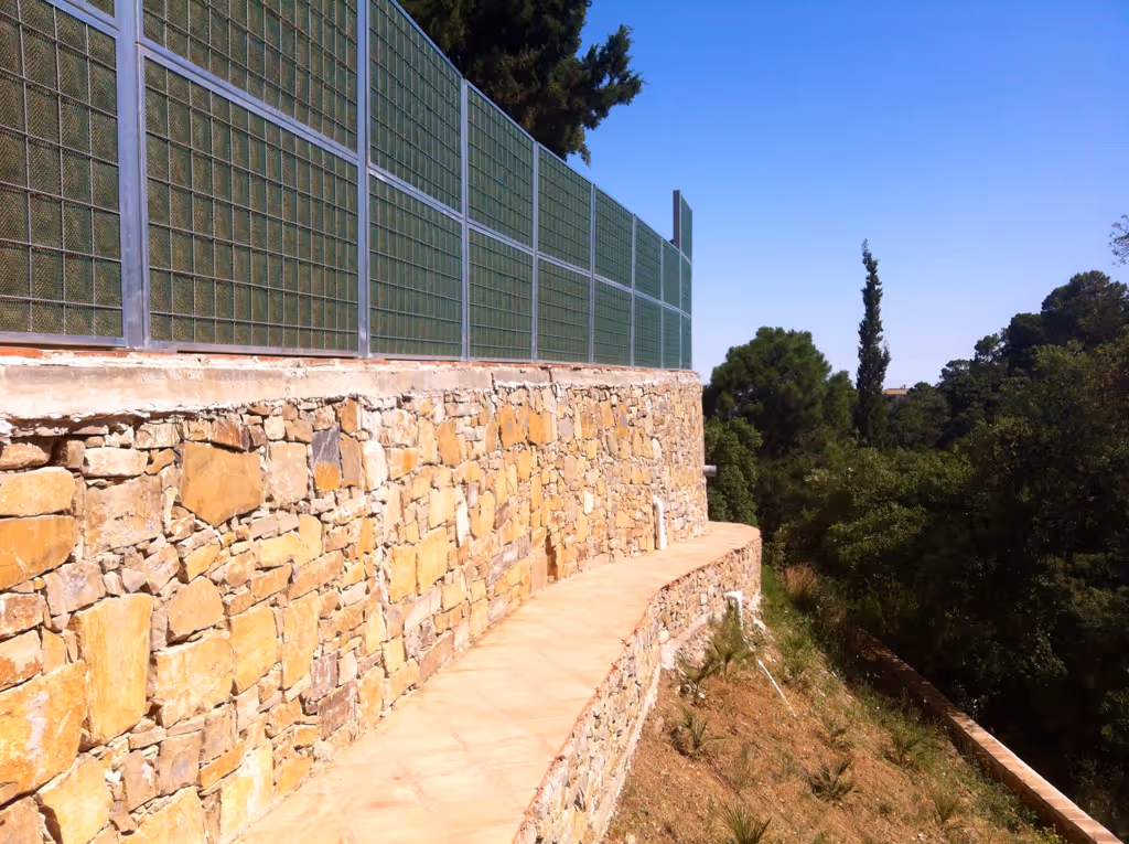 Stone retaining wall with a curved walkway and metal acoustic fence above, overlooking trees and clear sky.