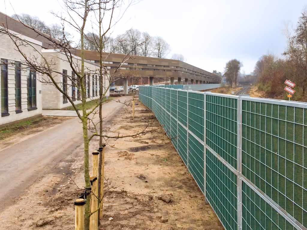 Une longue haie brise-bruit le long d'un chemin de terre, avec à proximité un bâtiment moderne et des arbres dépouillés.