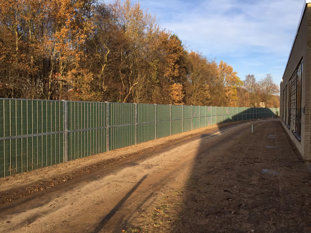 Une longue haie verte servant de brise-bruit longe un chemin de terre à côté d'un bâtiment et d'arbres aux couleurs d'automne.
