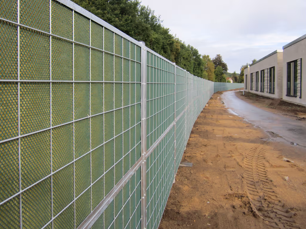 Une longue clôture acoustique métallique avec des panneaux verts longeant un chemin de terre, à côté d'un bâtiment moderne et d'arbres.