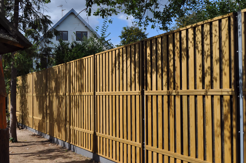 Long wooden acoustic fence beside a dirt path outside with a house and trees behind it.