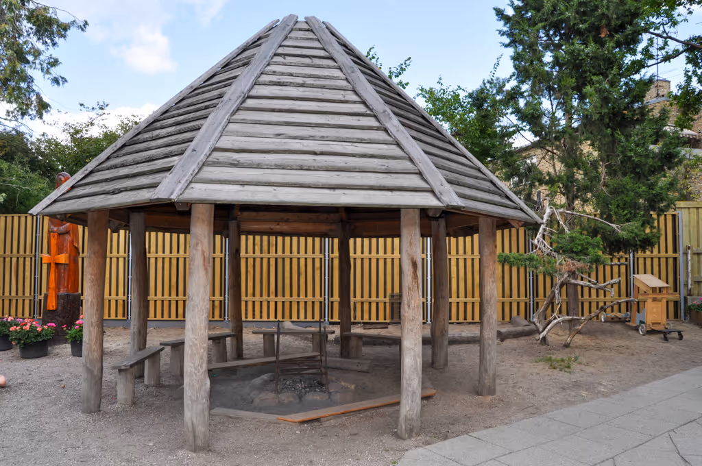 Wooden hexagonal gazebo with a pointed roof on a gravel surface next to an acoustic fence and trees.