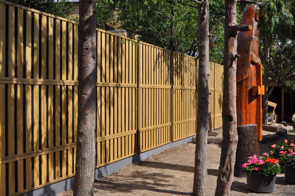 Tall wood acoustic fence with vertical slats beside trees and potted pink and red flowers in sunlight.