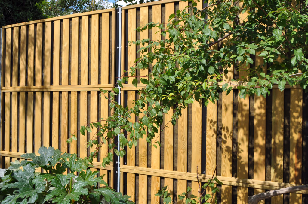 Wooden acoustic fence with vertical slats and plants with green leaves in front under sunlight.