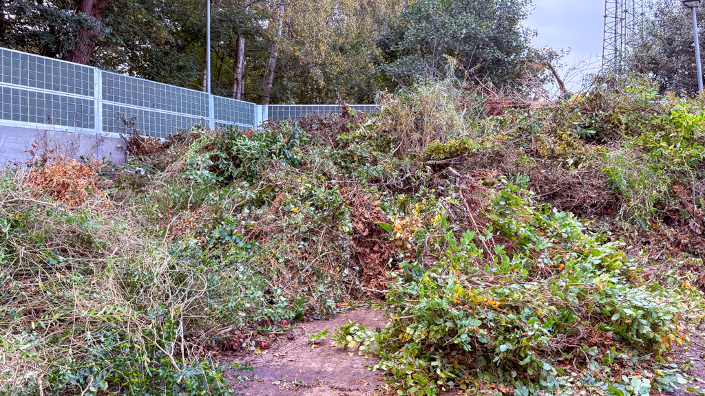 Large pile of green and brown garden waste in front of a steel acoustic fence with trees behind it.