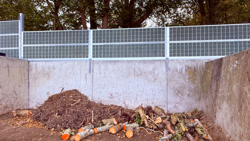 Piles of tree branches, logs, and twigs inside a concrete enclosure with a acoustic metal fence above it.