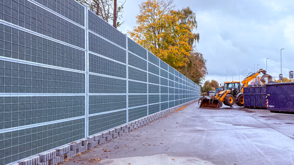 Long steel acoustic fence with grid panels near construction machinery and containers on a cloudy day.