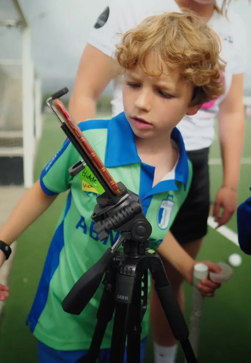 Jonge hockeyer kijkt nieuwsgierig naar een smartphone op een statief tijdens een training op het veld.