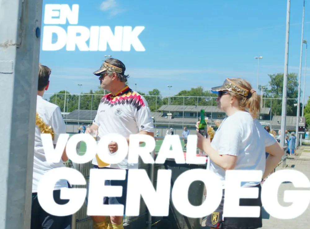 Twee mensen drinken bier langs een sportveld tijdens een zonnig toernooi, met grote tekst: 'EN DRINK VOORAL GENOEG'.