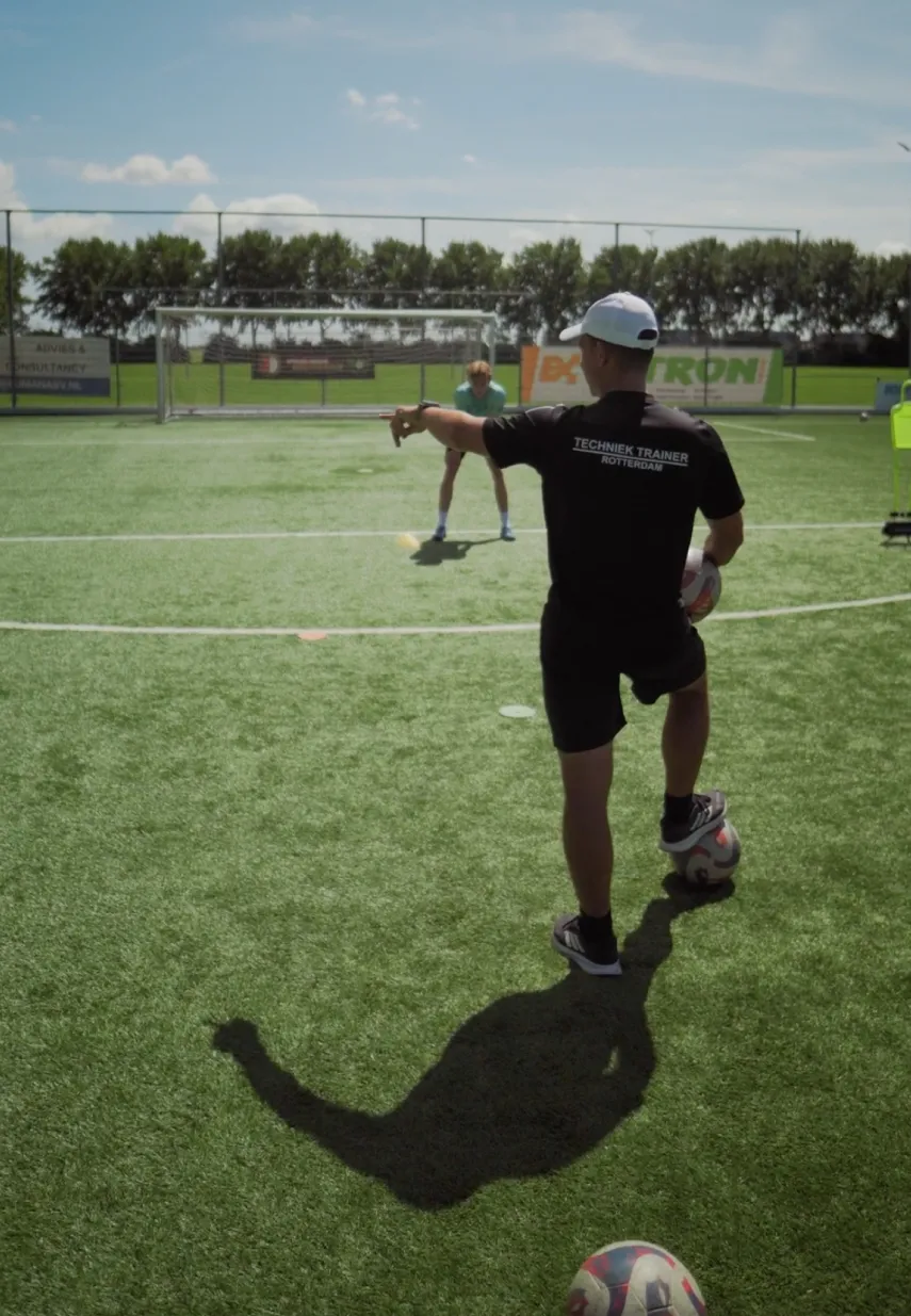 Voetbaltrainer wijst naar een speler op het veld tijdens een training, met 'Techniek Trainer Rotterdam' op zijn shirt.