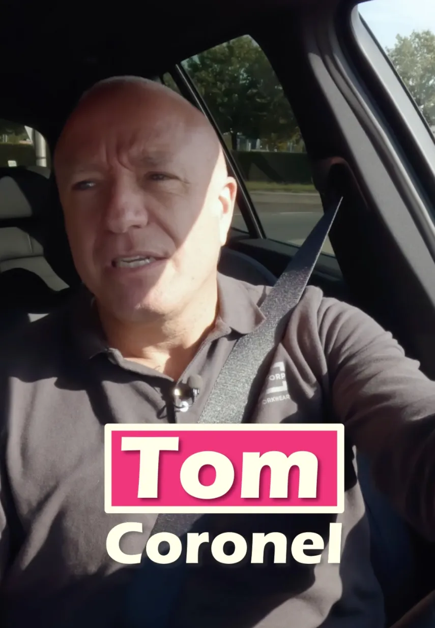 Tom Coronel zit pratend in een auto met gordel om, naam in roze-witte tekstkader in beeld.